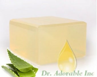 May include: A clear, rectangular block of soap base with a yellow drop of oil on top. The soap base is sitting on a white surface with a green aloe vera plant in the foreground. The text "Dr. Adorable Inc." is visible in the lower right corner of the image.