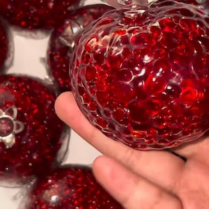 Op de afbeelding: Close-up van een groep rode glazen granaatappel ornamenten. De ornamenten zijn gemaakt van helder glas met een getextureerd oppervlak dat lijkt op de zaden van een granaatappel. De ornamenten zijn allemaal even groot en hebben dezelfde vorm.