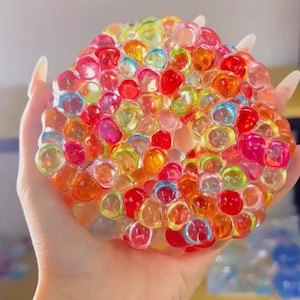 Op de afbeelding: Een kleurrijke, zachte bal gevuld met kleine, transparante, veelkleurige bolletjes. De bolletjes zijn in een willekeurig patroon gerangschikt en creëren een visueel aantrekkelijke, tactiele ervaring.