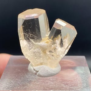 May include: A clear, faceted crystal cluster with a light amber hue, mounted on a clear acrylic block. The crystal has a natural, raw appearance, with visible internal structures. The base is a small, gray, rounded support.