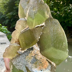 Op de afbeelding: Een grote, ruwe, geelgroene kristalgroep. Het kristal heeft een ruw, getextureerd oppervlak en wordt in de handen van een persoon gehouden. De achtergrond is wazig en toont groen gebladerte en een watermassa. Het kristal is een natuurlijk mineraal specimen.
