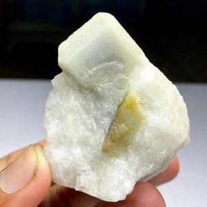 May include: A raw, uncut mineral specimen featuring a large, off-white crystalline structure with a rectangular top and a smaller, yellowish-green inclusion. The stone is held in a hand, showcasing its natural texture and color variations.