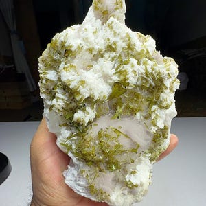 以下が含まれることがあります： 手で持たれた天然鉱物標本。標本は、緑と透明な結晶のクラスターを持つ白い基部が特徴です。 全体的な形状は不規則で質感があり、粗い自然な外観をしています。