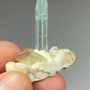 May include: A light blue aquamarine crystal stands upright on a base of white and tan minerals. The aquamarine is a clear, elongated prism with a pointed top. The base features several tan and white crystal formations. The background is a solid gray.