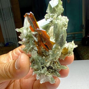 Puede incluir: Un grupo de cristales que presenta una mezcla de colores. Los tonos dominantes son verde claro y transparente, con toques de naranja y marrón. Los cristales tienen una textura rugosa y natural, sostenidos en una mano.