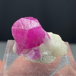 以下が含まれることがあります： 鮮やかなファセットルビーの結晶で、濃いピンク色をしています。白と灰色の岩を背景にしています。宝石の鋭いエッジと結晶構造がはっきりと見え、その自然な美しさを際立たせています。この標本は、鉱物芸術の印象的な例です。