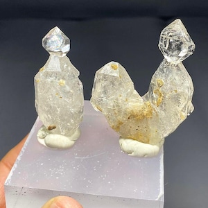 Puede incluir: Dos formaciones de cristales de cuarzo transparente con puntas, montadas sobre una base blanca. Los cristales tienen un aspecto translúcido con algunas inclusiones. La base es de color gris claro. Espécimen mineral natural.