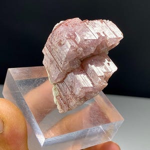Può includere: Un esemplare minerale grezzo rosa pallido con una struttura cristallina striata. Il gruppo presenta molteplici formazioni prismatiche verticali con una superficie traslucida che riflette la luce, montato su una base espositiva quadrata in acrilico trasparente.
