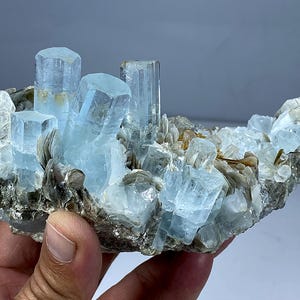 Może przedstawiać: Grupka jasnoniebieskich kryształów akwamarynu o krystalicznej strukturze osadzona na matrycy szarych i brązowych minerałów. Kryształy różnią się wielkością i kształtem, niektóre wyglądają jak wydłużone pryzmaty. Naturalny okaz geologiczny.