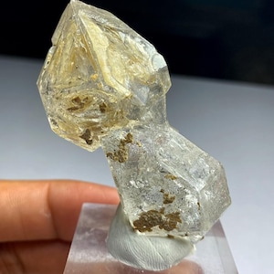 Puede incluir: Un cristal de cuarzo transparente con interior dorado e inclusiones marrones. El cristal está montado sobre una base acrílica transparente. El cuarzo tiene forma geométrica con bordes afilados y punta. Espécimen mineral natural.