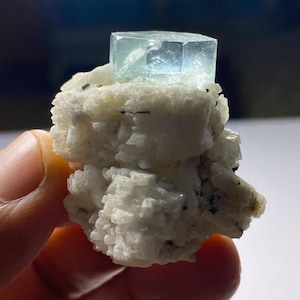Könnte beinhalten: Ein hellblaues Aquamarin-Kristall mit geometrischer Form sitzt auf einer weißen, strukturierten Mineralformation. Der Kristall ist durchscheinend, und die Mineralbasis hat ein raues, kristallines Aussehen. Kleine schwarze Einschlüsse sind sichtbar.