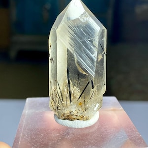 Puede incluir: Un cristal de cuarzo transparente con inclusiones de turmalina negra. El cristal está montado sobre una base rectangular transparente. El cuarzo tiene una punta y es transparente con líneas internas visibles e inclusiones negras en forma de aguja.