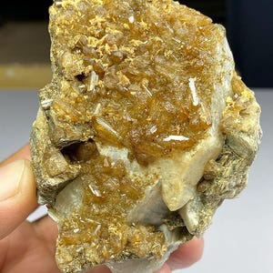 May include: A rough, natural mineral specimen featuring a mix of golden-brown and clear crystal formations. The crystals are clustered on a light-colored matrix, creating a textured appearance. The overall color palette is earthy, with variations in transparency.