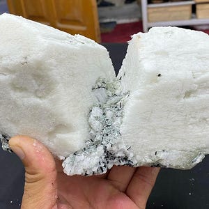 May include: A large, white mineral specimen with a rough, textured surface. The stone is bisected, revealing a crystalline structure with dark inclusions. The mineral is held in a hand, showcasing its size and detail.