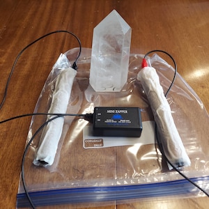 May include: A clear quartz crystal stands between two white-wrapped cylinders connected by black wires to a black box labeled "Mini Zapper." The box is inside a clear plastic bag on a wooden surface.