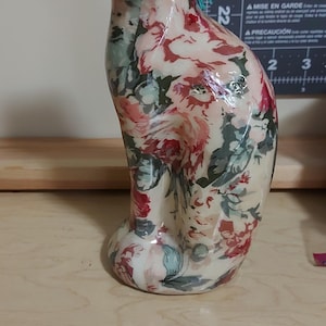 May include: A ceramic cat ornament featuring a floral design in pink, red, and green hues. The cat has a blue collar and is in a seated position. The ornament is placed on a wooden surface.