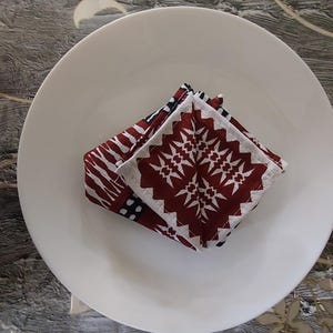 May include: A folded square napkin with a geometric pattern in burgundy and white, resting on a white ceramic plate. The napkin's design features a repeating starburst motif. The plate is centered on a textured surface with floral accents.