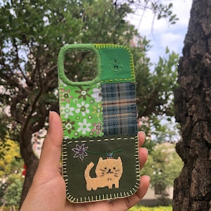 Puede incluir: Funda de teléfono patchwork hecha a mano con estampado floral verde, cuadros escoceses y una aplicación de gato. La funda tiene un borde verde y un diseño cosido. La funda del teléfono se sostiene frente a un fondo verde borroso.