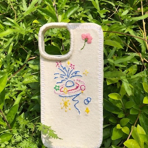 May include: A cream-colored canvas phone case featuring hand-embroidered designs. The embroidery includes a small pink flower, a blue and pink planet with rings, and various yellow and blue stars and abstract shapes. The edges are stitched in white.
