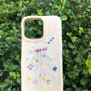 Puede incluir: Una funda de teléfono de color crema con detalles bordados a mano. El diseño incluye una medusa central en tonos rosa, amarillo y azul, rodeada de pequeñas flores, corazones y una letra B bordados. Los bordes están rematados con costuras.
