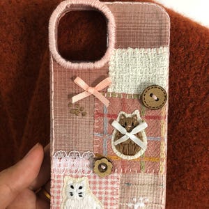 May include: A phone case featuring a patchwork design of pink and white fabric squares with embroidered details. It includes a white cat, a brown cat, two wooden buttons, a small pink ribbon bow, a white lace trim, and a paw print motif.