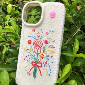 May include: A rectangular phone case made of off-white canvas fabric featuring hand-embroidered floral designs. The embroidery includes a central red rose bouquet with a bow, surrounded by small colorful flowers, stars, and hearts in red, blue, and yellow.