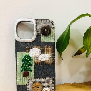 Pode incluir: Uma capa de telemóvel em ganga cinzenta escura com remendos bordados de um pinheiro verde, nuvens brancas e chuva azul. A capa é decorada com três botões circulares em castanho e branco, com costura branca à volta da abertura da câmara.