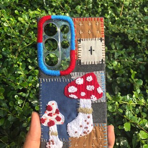 Può includere: Una custodia per telefono rettangolare con un design patchwork di quadrati in denim e tessuto con ricami fatti a mano. La custodia mostra due funghi ricamati con cappelli rossi e macchie bianche, accentuati da toppe in tessuto blu e marrone.