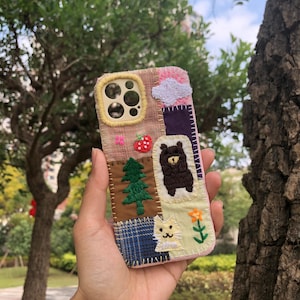 May include: A patchwork phone case featuring embroidered motifs of a bear, cat, strawberry, tree, and cloud. The case has a pink border and a yellow camera cutout. The case is held in front of a blurred background of trees.