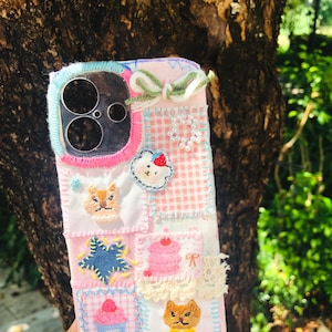Op de afbeelding: Een rechthoekig telefoonhoesje met een patchworkontwerp van lichtroze en witte stoffen vierkantjes. Het hoesje is versierd met geborduurde motieven, waaronder een ster, een cupcake en twee kattengezichten, samen met een klein garenstrikje en kant.