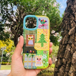 Puede incluir: Una funda de teléfono patchwork con un borde azul y varios diseños bordados. La funda presenta un gato, flores y un árbol de Navidad. La funda se sostiene frente a un fondo verde borroso.