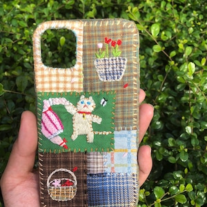 Könnte beinhalten: Bestickte Patchwork-Handyhülle mit einer skurrilen Katze, Blumen und einem Korb. Die Hülle besteht aus verschiedenen Stoffmustern, darunter Karos und Vichy, mit einem genähten Rand. Das Design beinhaltet rote, rosa, grüne und braune Farben.