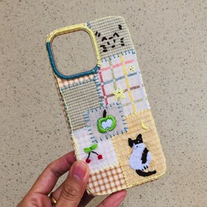 Könnte beinhalten: Eine rechteckige Handyhülle mit einem Patchwork-Design aus bestickten Stoffquadraten in Gelb, Weiß und Blau. Die Hülle enthält gestickte Motive eines grünen Apfels, roter Kirschen, eines gelben Sterns, einer Mondsichel sowie einer schwarz-weißen Katze.