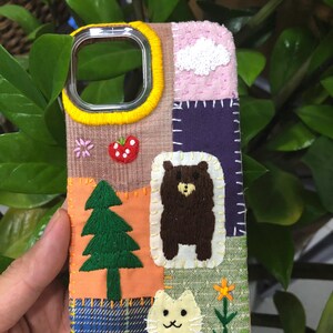 May include: A phone case featuring a patchwork design of various fabric textures and colors, decorated with hand-embroidered motifs including a brown bear, a white cat, a green pine tree, a red strawberry, a white cloud, and a yellow flower.