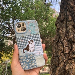 Op de afbeelding: Een patchwork telefoonhoesje met een kattenborduurwerk. De hoes heeft een lichtblauw en wit kleurenschema met verschillende stofpatronen, waaronder ruitjes en bloemmotieven. De kat is bruin en wit.