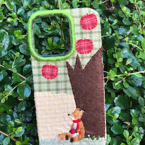 May include: A rectangular phone case featuring a fabric cover with a green and white plaid pattern. The design includes a brown felt tree, three red circular fabric patches, and an embroidered fox wearing a red scarf sitting on the ground.