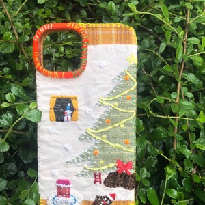 May include: A rectangular phone case featuring white fabric with embroidered Christmas motifs including a green tree, a snowman in a window, a red bow, a gingerbread man, and a candle. The edges are stitched with orange and yellow thread.