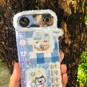 May include: A blue and white phone case featuring a patchwork design of gingham and floral fabric. The case is decorated with a white yarn border, a frayed fabric bow, a small white button, a butterfly charm, lace trim, and a patch with a cat illustration.