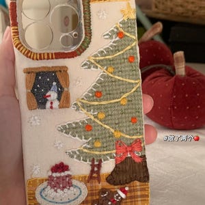 Op de afbeelding: Geborduurde telefoonhoes met een kerstthema. Het ontwerp omvat een kerstboom, een sneeuwpop en een peperkoekmannetje. De hoes heeft een cr&egrave;mekleurige achtergrond met een ruitaccent en rode en groene randen. Inclusief een ster, ornamenten en een rode strik.