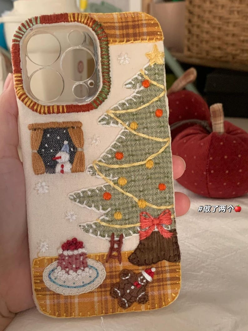 Op de afbeelding: Geborduurde telefoonhoes met een kerstthema. Het ontwerp omvat een kerstboom, een sneeuwpop en een peperkoekmannetje. De hoes heeft een cr&egrave;mekleurige achtergrond met een ruitaccent en rode en groene randen. Inclusief een ster, ornamenten en een rode strik.