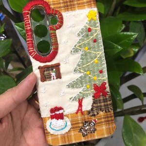 May include: A rectangular phone case featuring a cream-colored fabric center with hand-embroidered Christmas motifs including a green tree, a snowman in a window, and a gingerbread man. The edges are trimmed with a yellow and brown plaid fabric.