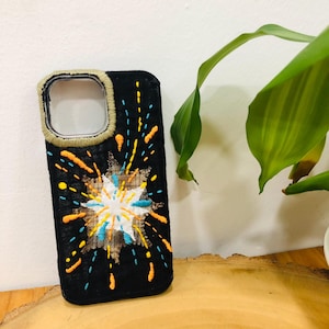 May include: A black phone case featuring hand-embroidered fireworks in white, bright blue, and orange thread. The design includes a central starburst pattern with radiating lines and dots. The camera cutout is bordered by a thick, tan-coloured stitched edge.