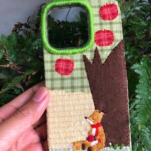 Może przedstawiać: Haftowane etui na telefon z fantazyjną sceną leśną. Etui ma zielono-biały wzór w kratkę, czerwone akcenty jabłkowe i brązowe drzewo. Na dole wyhaftowano lisa z czerwoną szarfą. Obramowanie aparatu w etui jest zielone.