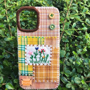 May include: A rectangular phone case covered in patchwork fabric featuring plaid patterns in yellow, orange, and brown. It includes a white embroidered patch with colorful flowers, two small round buttons, and a small orange tag labeled Hand Made.