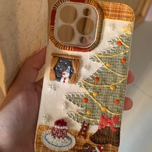 Op de afbeelding: Geborduurde telefoonhoes met een kerstontwerp. De hoes heeft een kerstboom, sneeuwpop en peperkoekmannetje. De achtergrond is cr&egrave;mekleurig met een geruite rand en rode en groene afwerking. Inclusief een kleine taart en een ladder.