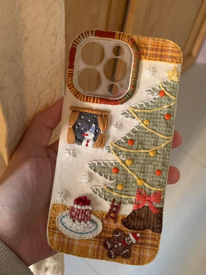 Op de afbeelding: Geborduurde telefoonhoes met een kerstontwerp. De hoes heeft een kerstboom, sneeuwpop en peperkoekmannetje. De achtergrond is cr&egrave;mekleurig met een geruite rand en rode en groene afwerking. Inclusief een kleine taart en een ladder.
