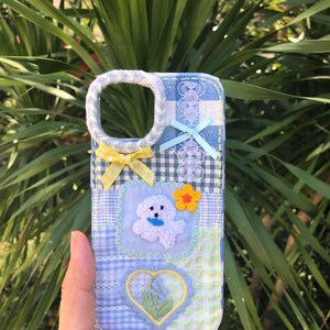 May include: A rectangular phone case featuring a blue and white chequered fabric patchwork design. It is decorated with a braided border around the camera cutout, two small fabric bows, a white embroidered dog patch, a yellow flower, and a heart patch.