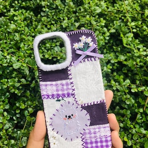 May include: A patchwork phone case in shades of purple, white, and lavender. The case features a floral design, a small bow, and a cute cartoon dog. The case is made of fabric and has a white border around the camera opening.