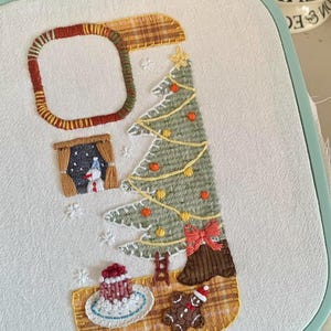 Op de afbeelding: Handgestikte kerstborduurwerk op witte stof, gezet in een mintgroene hoepel. Voorzien van een kerstboom met ornamenten, een raam met een sneeuwpop, een peperkoekmannetje en een cake. Het ontwerp bevat geruite en andere textuurstoffen.