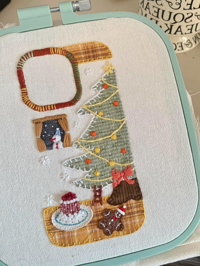 Op de afbeelding: Handgestikte kerstborduurwerk op witte stof, gezet in een mintgroene hoepel. Voorzien van een kerstboom met ornamenten, een raam met een sneeuwpop, een peperkoekmannetje en een cake. Het ontwerp bevat geruite en andere textuurstoffen.
